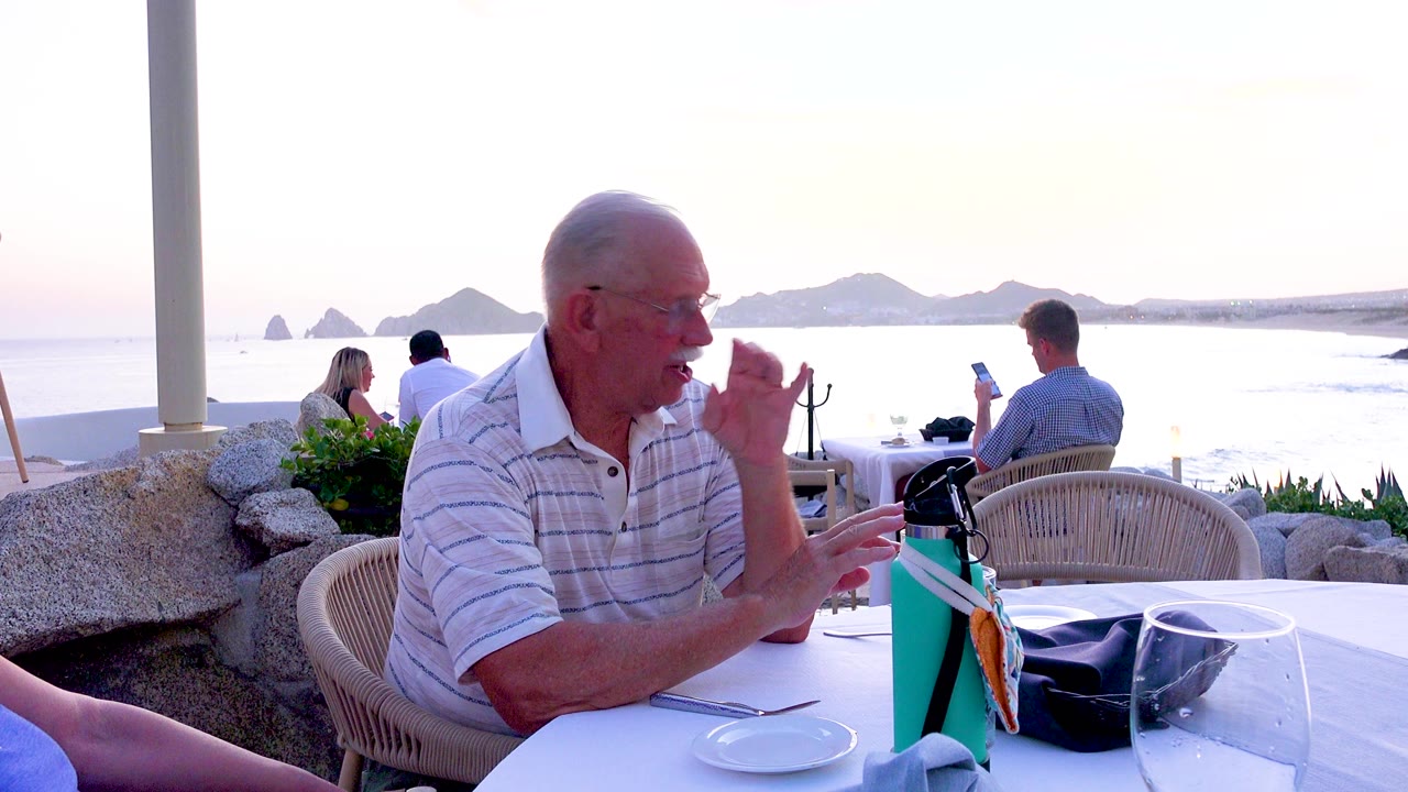 Senior man continues gesturing beside a white table with ocean behind him.