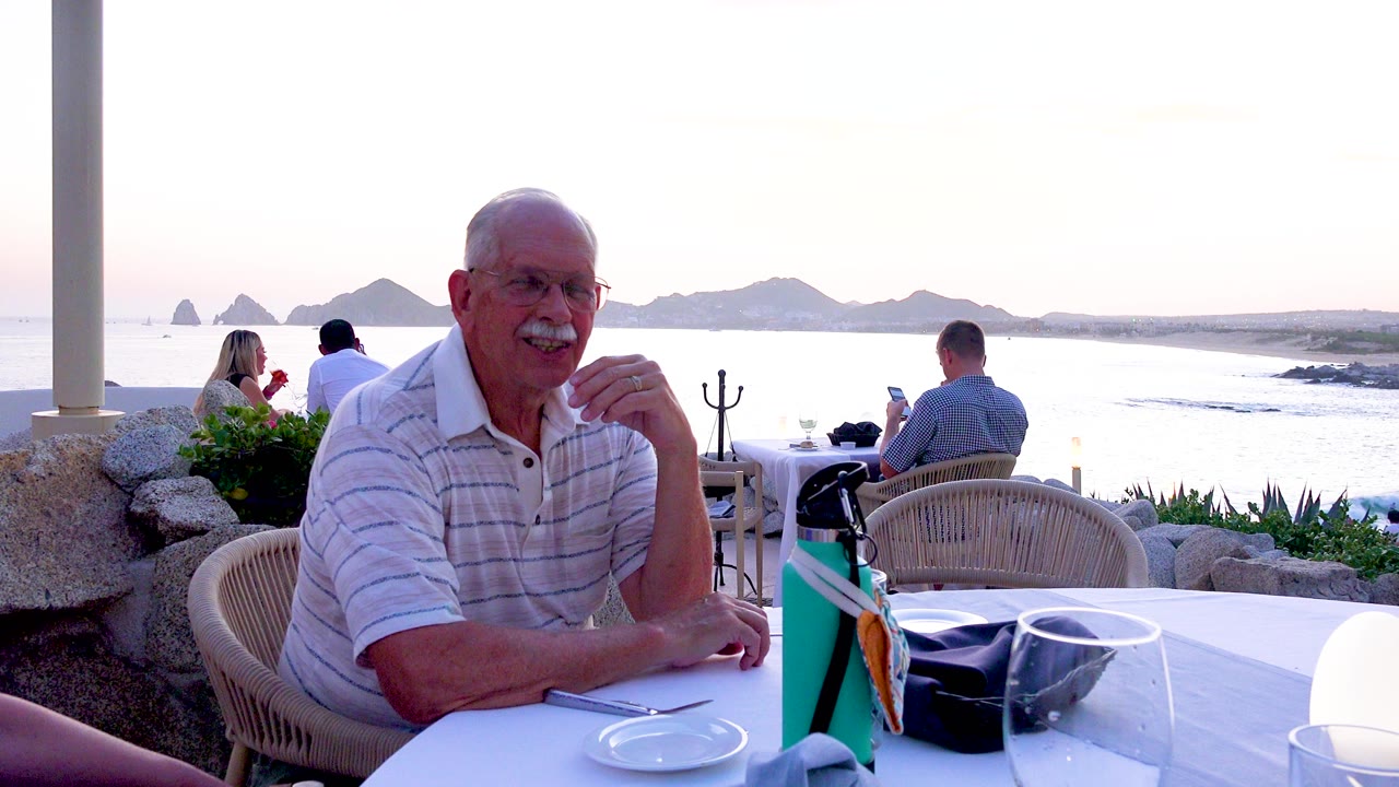 Senior man with glasses sits at a white outdoor restaurant table, with other diners, bright ocean water, and distant islands visible behind him.
