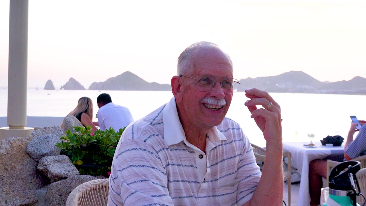 Senior man with glasses and a white mustache laughs at a patio table while background diners sit near a stone wall and hazy islands appear across the ocean.