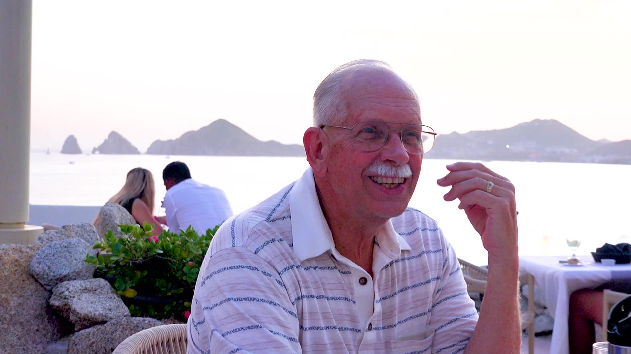 A closer framing catches the senior man laughing toward the side of the table while background diners face the water and dark island shapes sit on the bright horizon.
