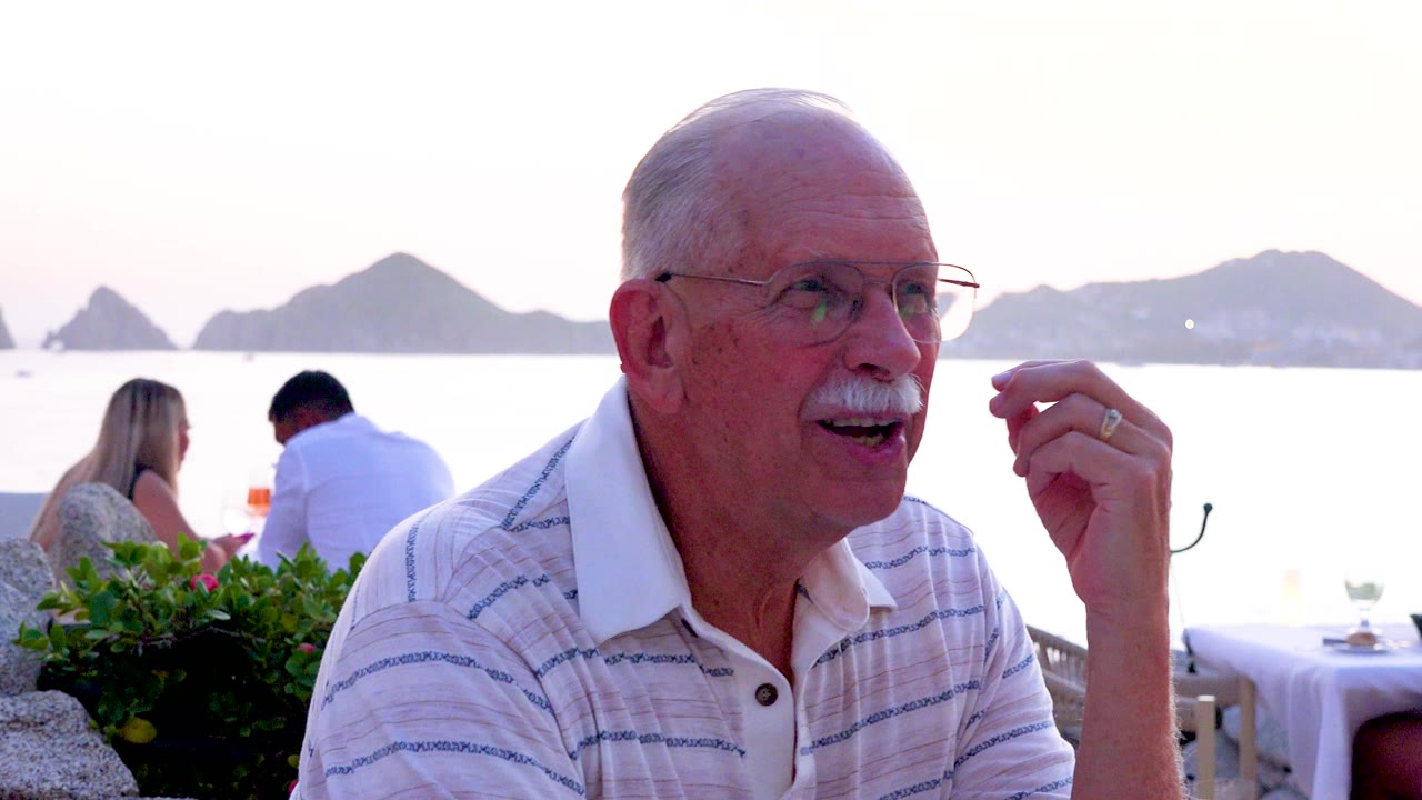 Senior man looks off to the side with his hand raised, with seated diners, white restaurant tables, calm water, and mountain silhouettes in the background.