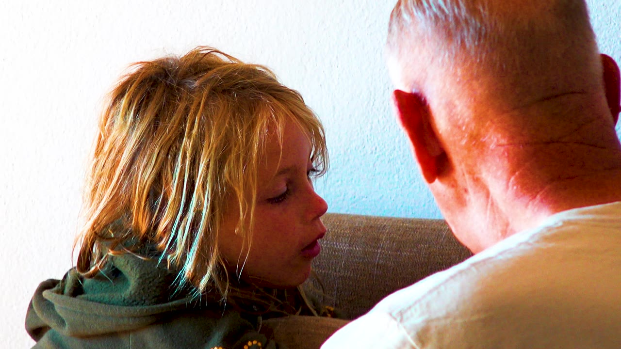 A blonde girl in a green hoodie sits in profile on a couch beside an older man seen from behind in a white shirt against a plain indoor wall.