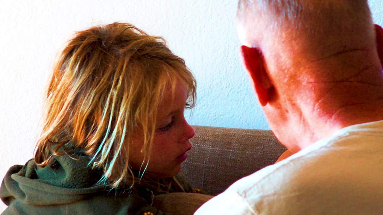The child leans slightly forward on the couch, still facing the man whose head and shoulder stay in the right side of the close indoor composition.