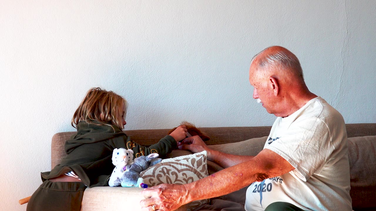 Child leans across sofa cushions while senior man watches and reaches forward.