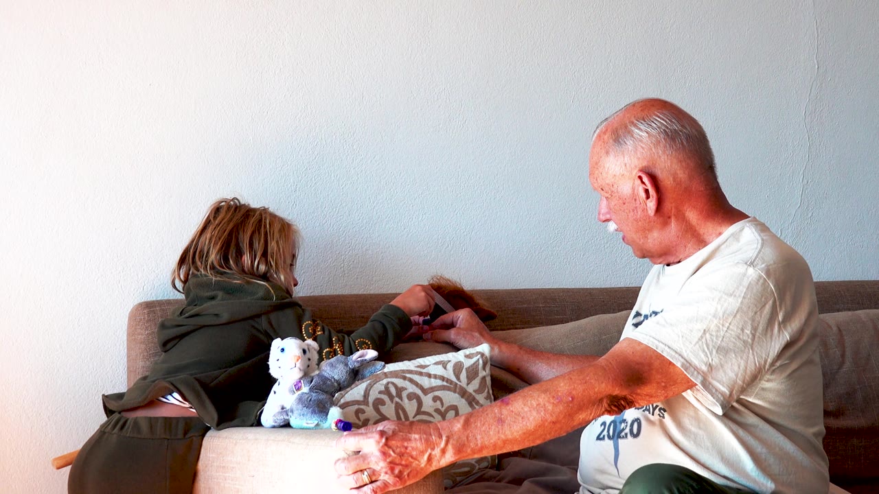 The child extends an arm toward the cushions near the ottoman while the senior man sits close by, facing the play area in a bright living room.