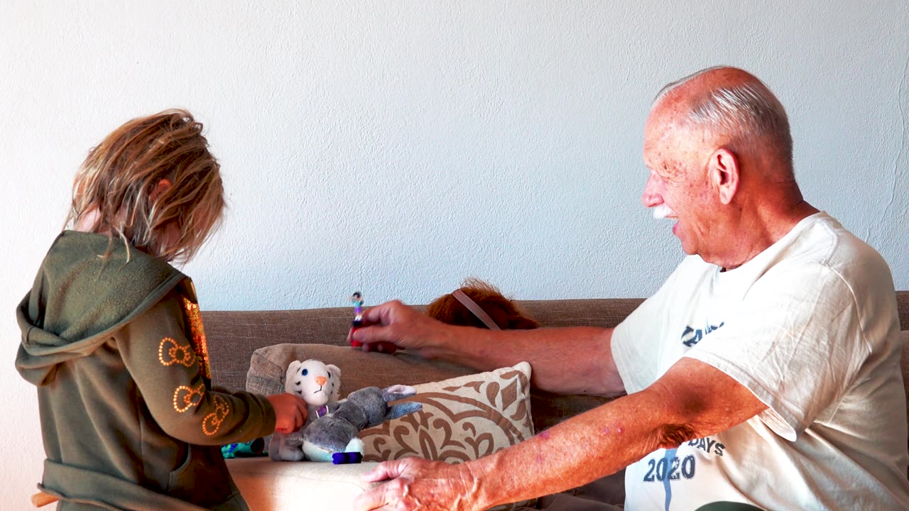 The older man extends his arm toward a small toy figure on the coffee table while the child stands close to stuffed animals in front of the sofa.