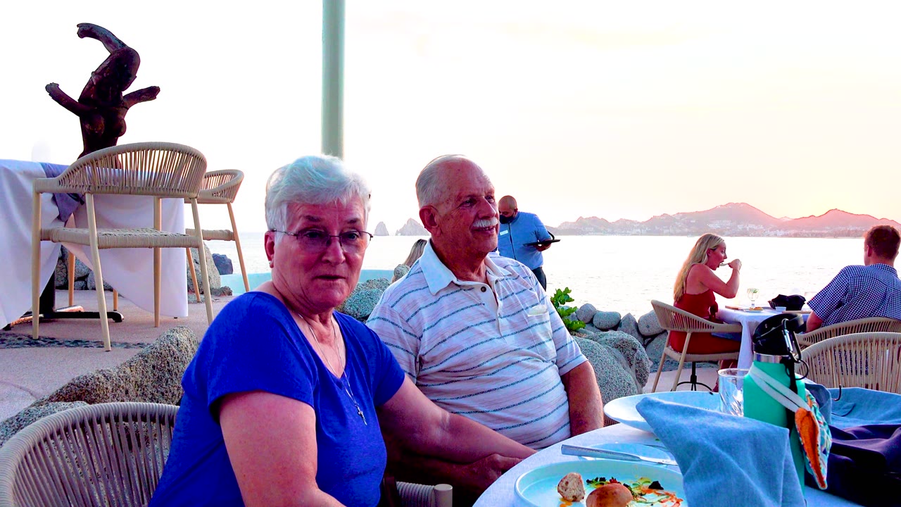 Senior couple beside table with waterfront diners in background.