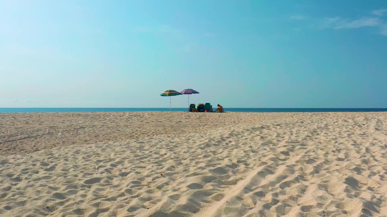 High-angle view of Desert Park Natural Reserve beach in Baja California Sur, with rippled sand in front, two umbrellas and distant people near the shoreline, and blue ocean beyond.