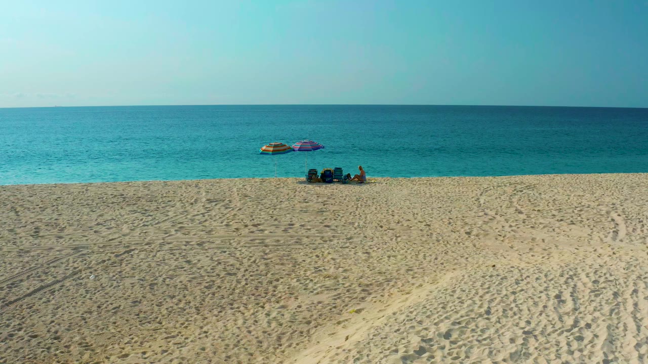 Opening aerial frame over Desert Park Natural Reserve in Baja California Sur, Mexico, with textured sand leading to two umbrellas, a small seated group, turquoise ocean, and blue sky.
