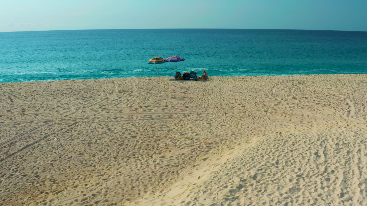 High aerial frame from Desert Park Natural Reserve in Baja California Sur showing textured sand, tire tracks, turquoise ocean, umbrellas, chairs or gear, and distant visible beachgoers near the shoreline.