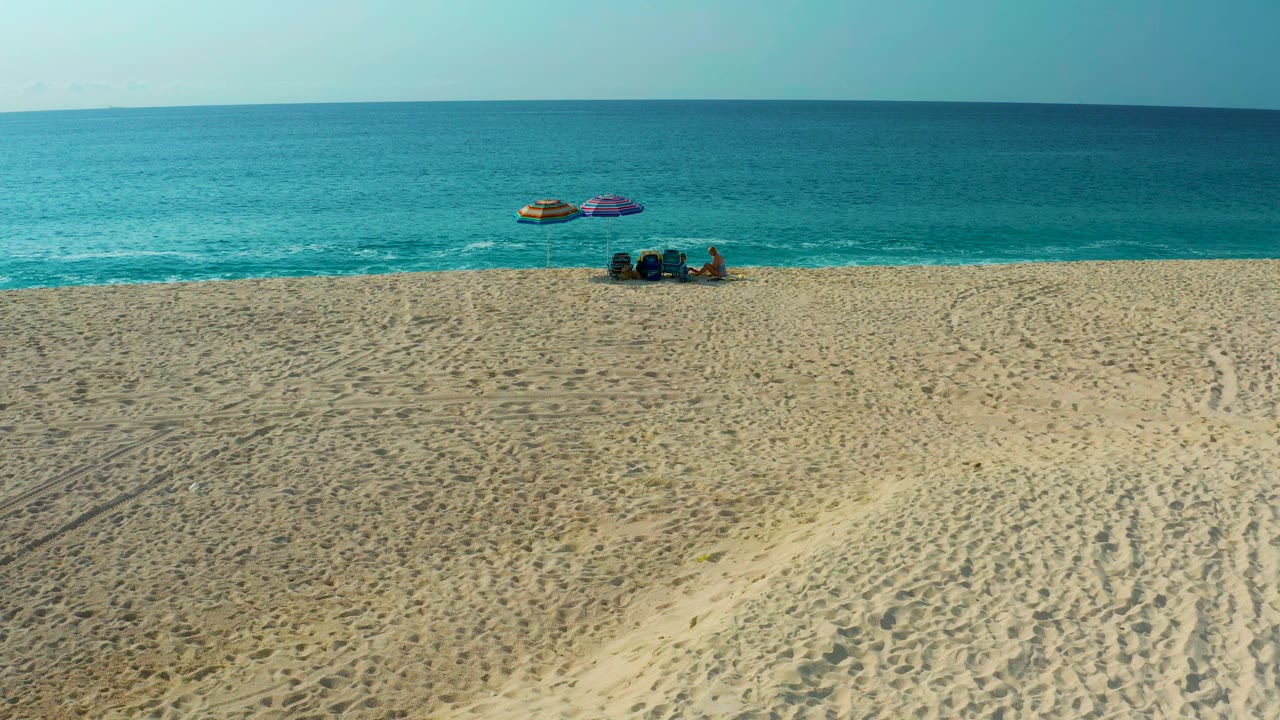 Aerial view in Baja California Sur with the beach setup slightly left of center, pale sand in the foreground, small wave foam at the shoreline, and visible people beside colorful umbrellas.