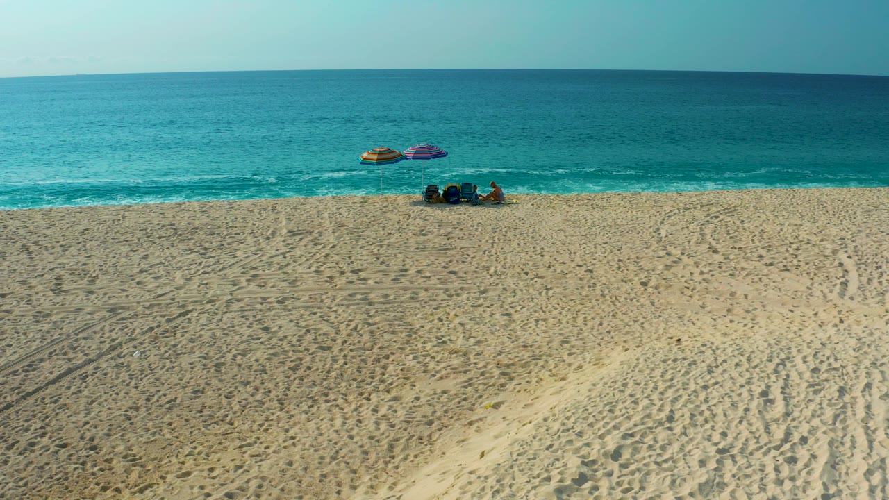 Drone-like high-angle frame from Desert Park Natural Reserve showing a broad empty stretch of rippled sand, a blue-green ocean horizon, umbrellas, beach gear, and a small group of visible people near the waterline.