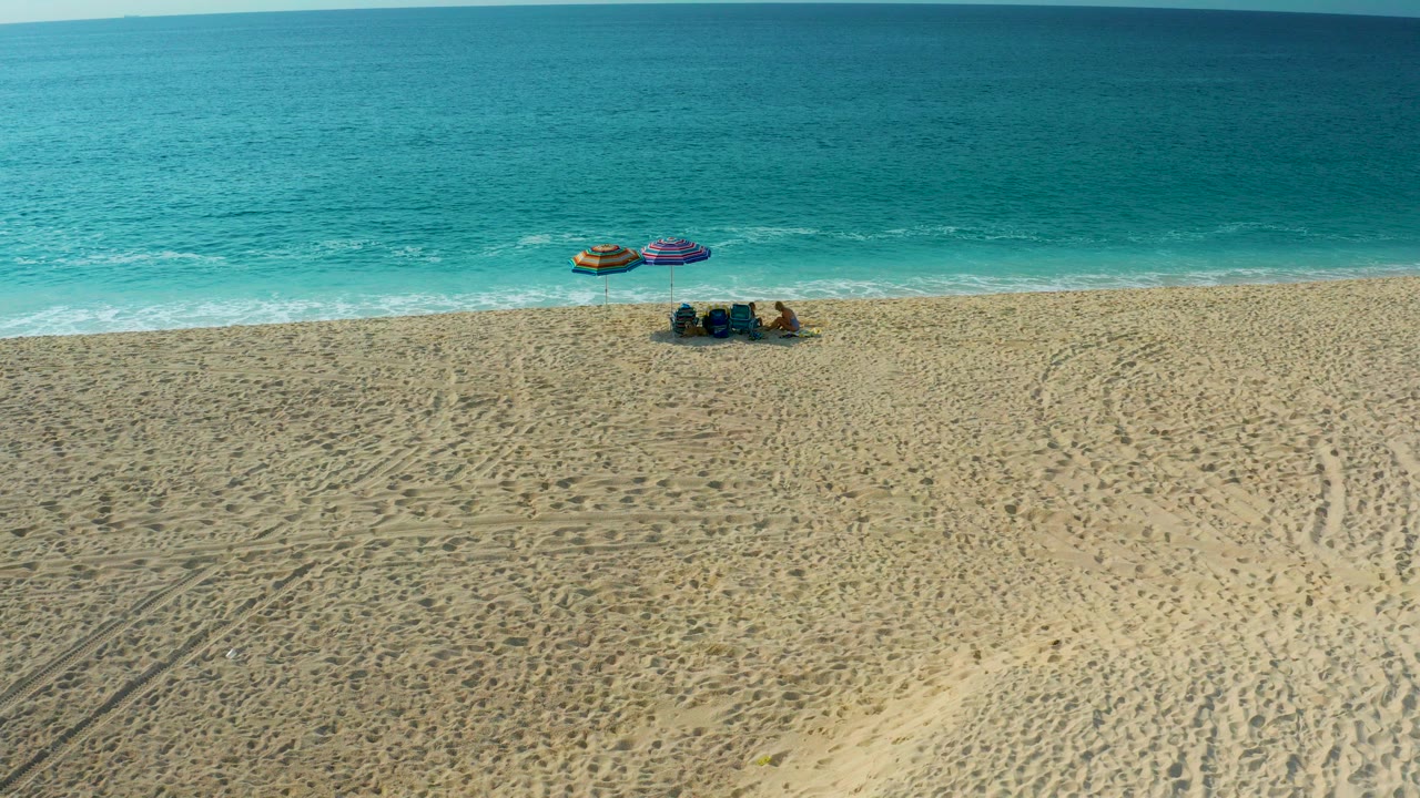 High-angle aerial frame from Desert Park Natural Reserve in Baja California Sur showing a small group on golden sand near two umbrellas, with turquoise ocean and a thin surf line behind them.