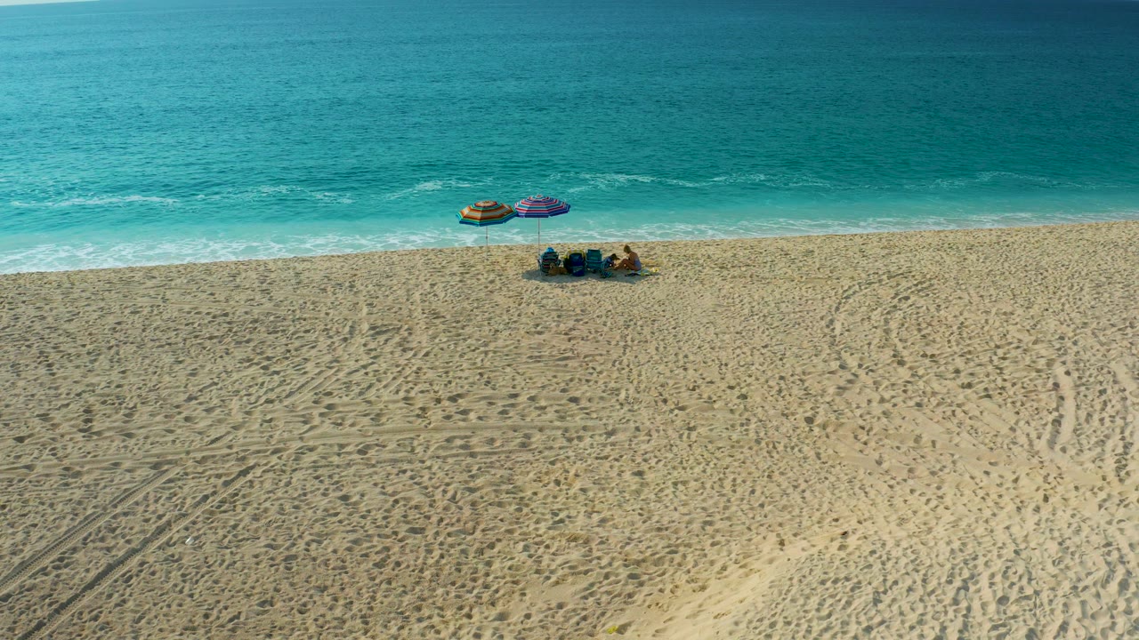 High oblique view over Desert Park Natural Reserve beach, where two umbrellas and a seated group sit below the turquoise ocean band and above tire tracks in the foreground sand.