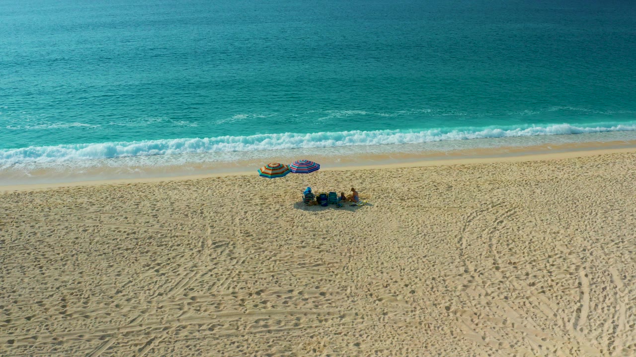 Opening aerial frame over Desert Park Natural Reserve in Baja California Sur, Mexico, showing visible beachgoers beside purple and yellow umbrellas below a band of turquoise surf.