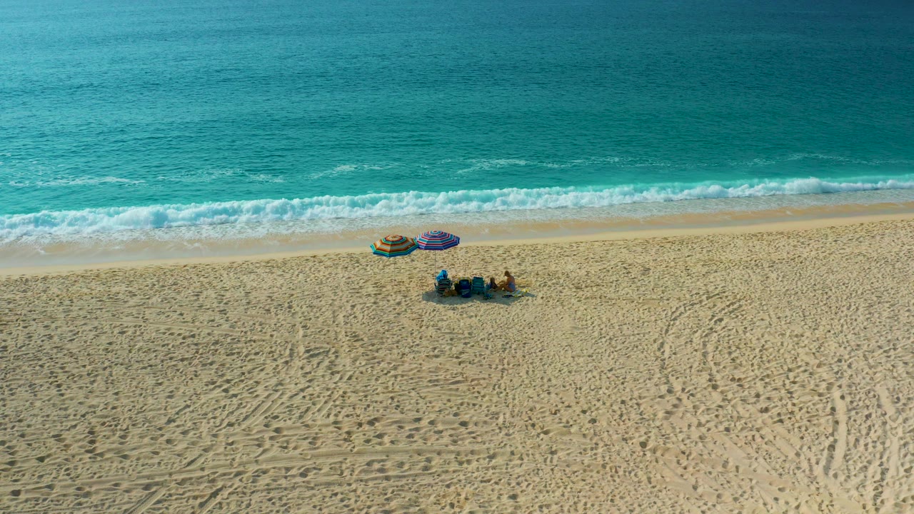 Beachgoers and two umbrellas on wide sand below a strip of breaking surf.