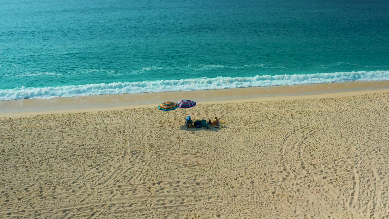 High-angle frame over Baja California Sur sand showing the beachgoers near two umbrellas, with the surf band set farther up the frame and textured tracks visible across the beach.
