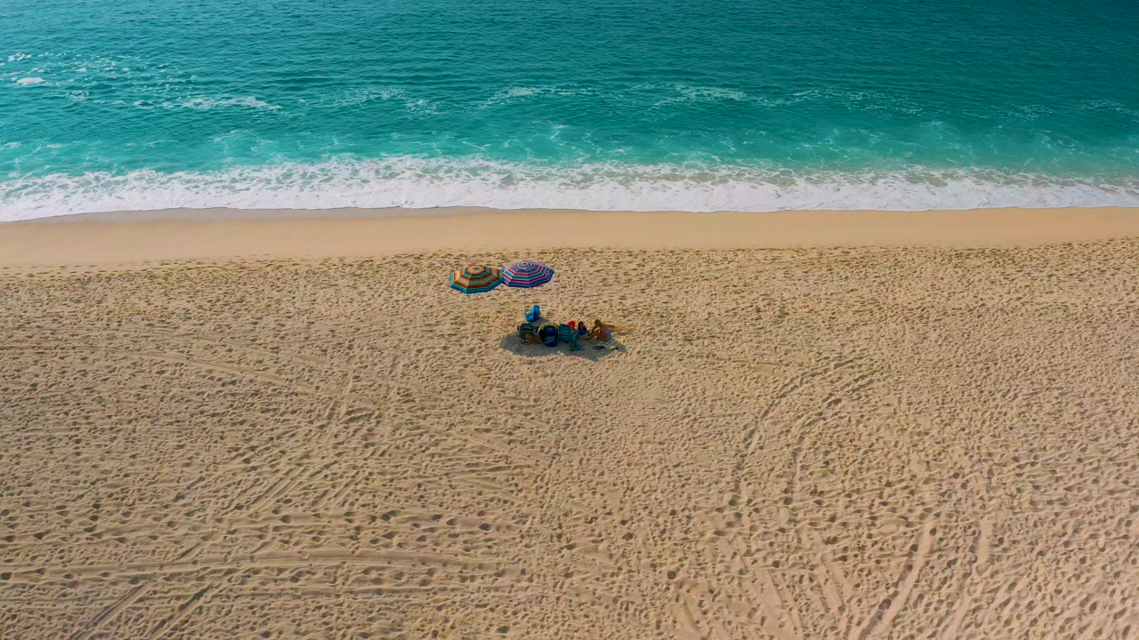Aerial view of waves, empty sand, tire marks, two umbrellas, and people relaxing on the beach.