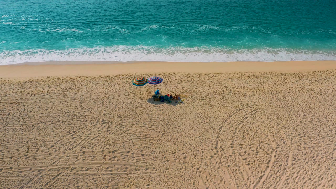 High overhead view in Desert Park Natural Reserve with a wider stretch of empty sand to the right of the visible beach group, turquoise ocean above, foam at the waterline, and tire tracks crossing the lower sand.