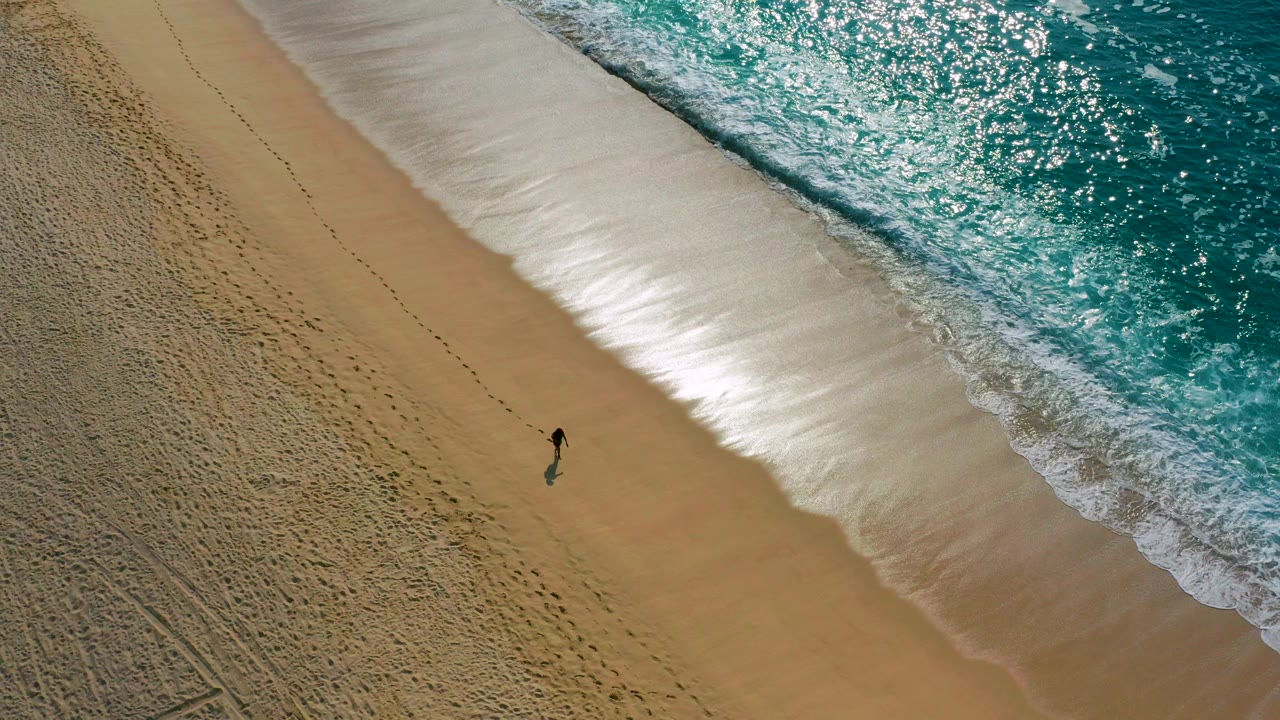 High-angle aerial frame from Desert Park Natural Reserve, Baja California Sur, showing one distant person on tan sand with white surf and bright turquoise water along the right edge.