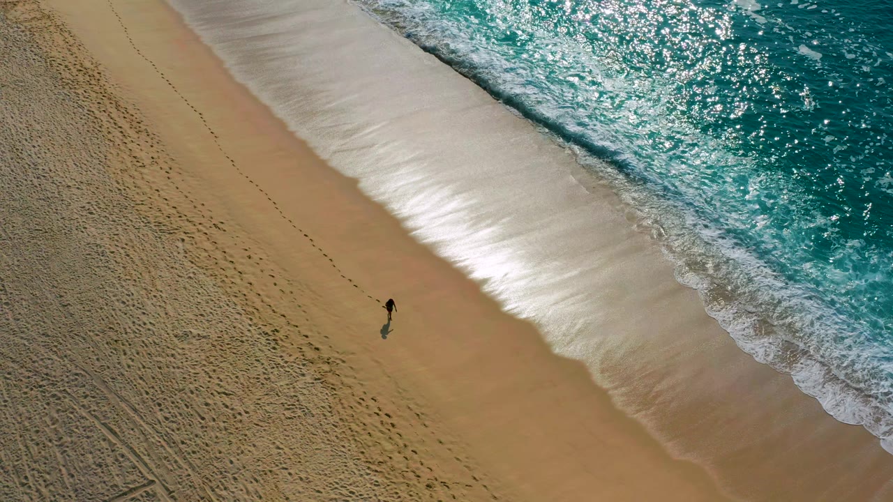 Top-down beach view with a solitary walker near the surf line.