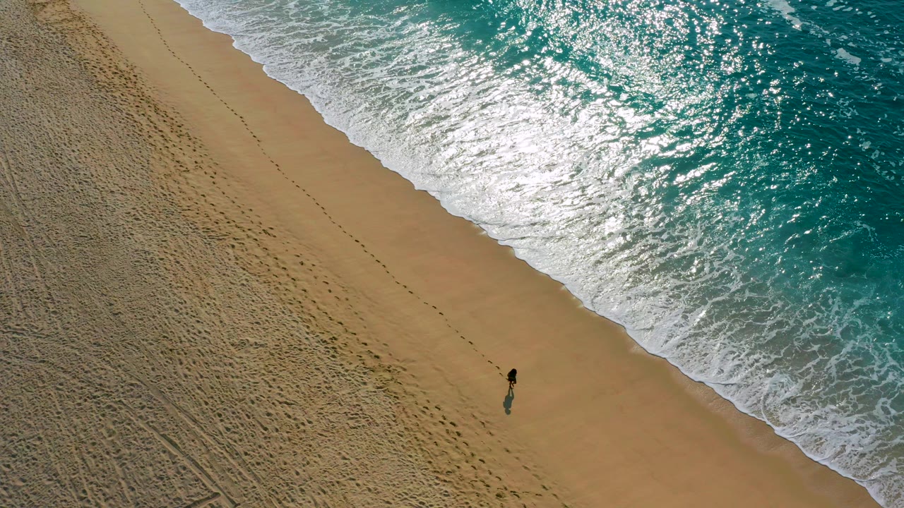 High-angle aerial frame in Baja California Sur, Mexico, showing one distant person on tan sand near a diagonal edge of white foam and turquoise ocean.