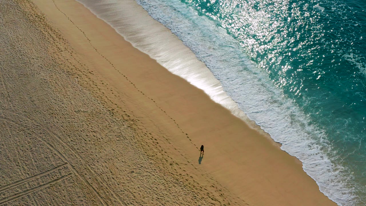 Overhead view in Desert Park Natural Reserve, Baja California Sur, with one small person walking on tan sand beside turquoise surf and sunlit water.