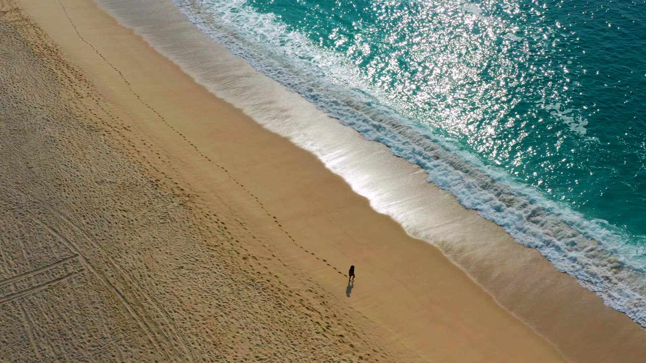 High aerial frame from Desert Park Natural Reserve in Baja California Sur shows a lone person on open sand below a band of white surf and glittering turquoise water.