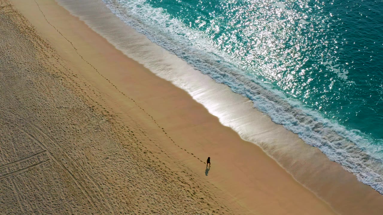 Person walking along a sandy shoreline with footprints and breaking waves.
