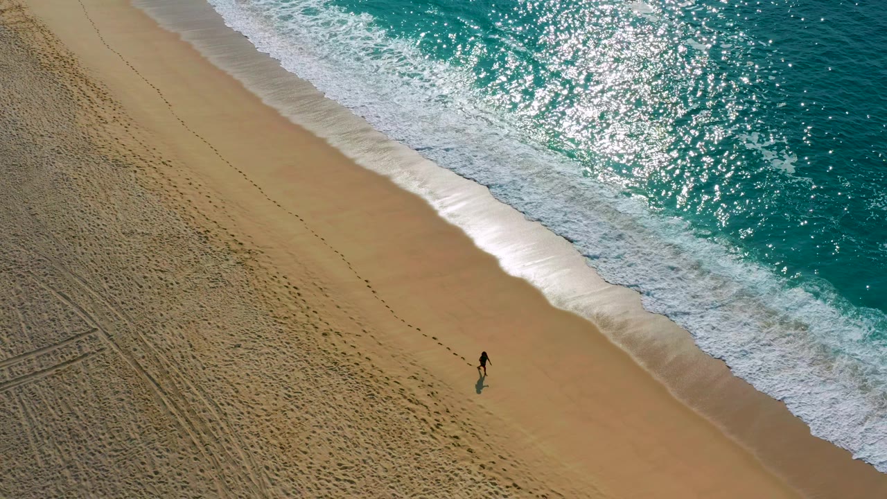 The frame shows the person closer to the waterline in a high-angle beach view, with white foam spreading across the upper shore and open sand around them.