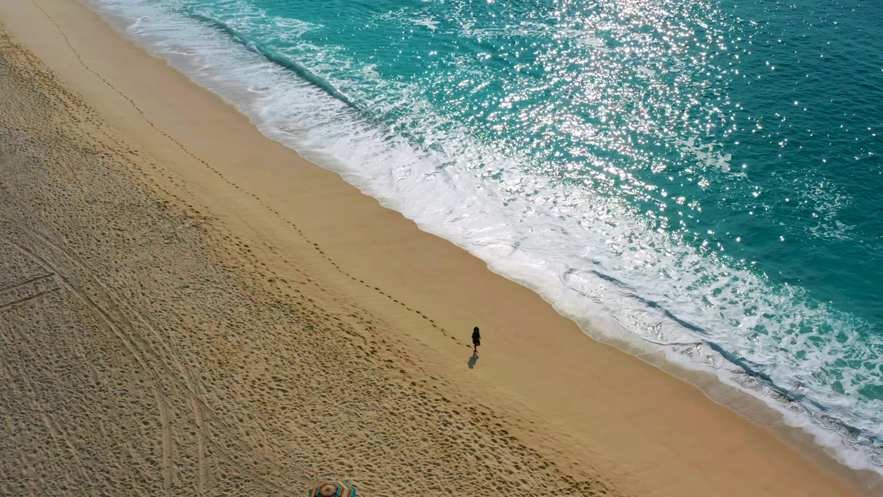 Distant person on sandy shoreline below sparkling ocean water.