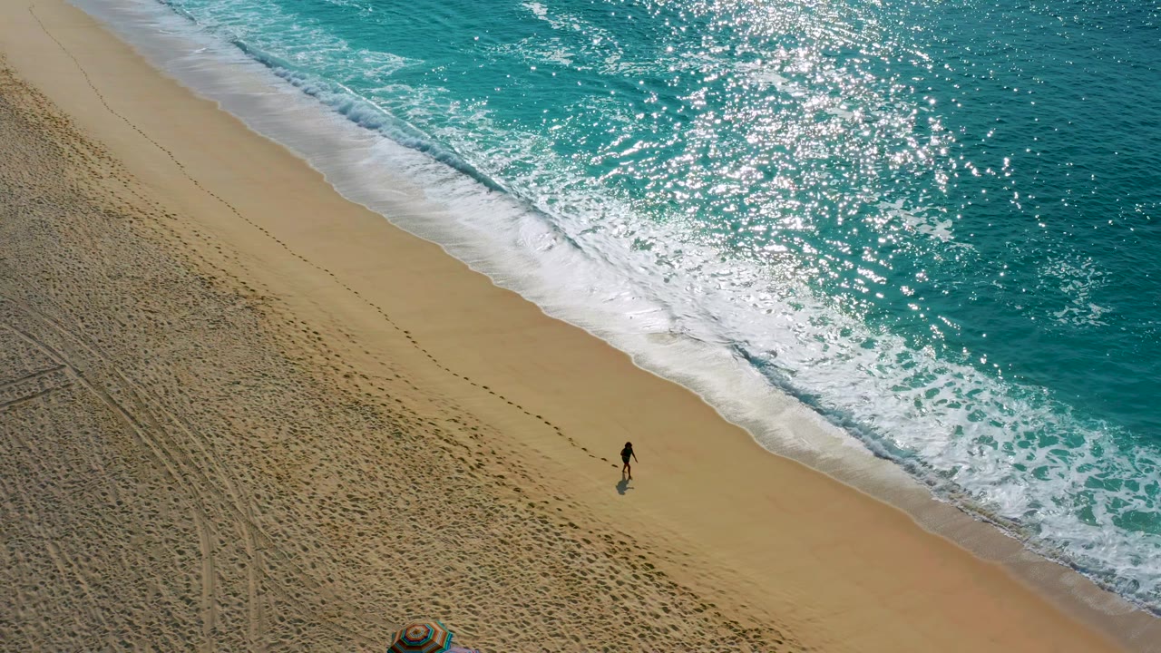 Drone-height frame in Baja California Sur showing the lone figure near the wet sand while white foam spreads along the turquoise shoreline.