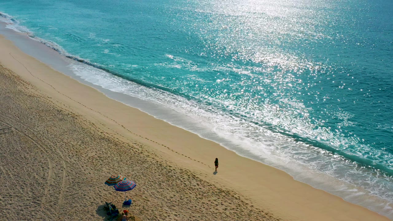 Drone view of a sandy shoreline with a small walker and beach umbrella below bright ocean glare.