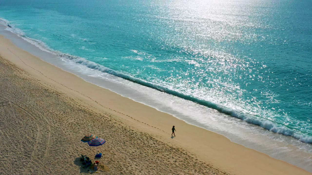 High-angle view in Baja California Sur, Mexico, showing the person farther along the shoreline, a breaking wave near the upper right, and the blue umbrella and chairs set back on the sand.