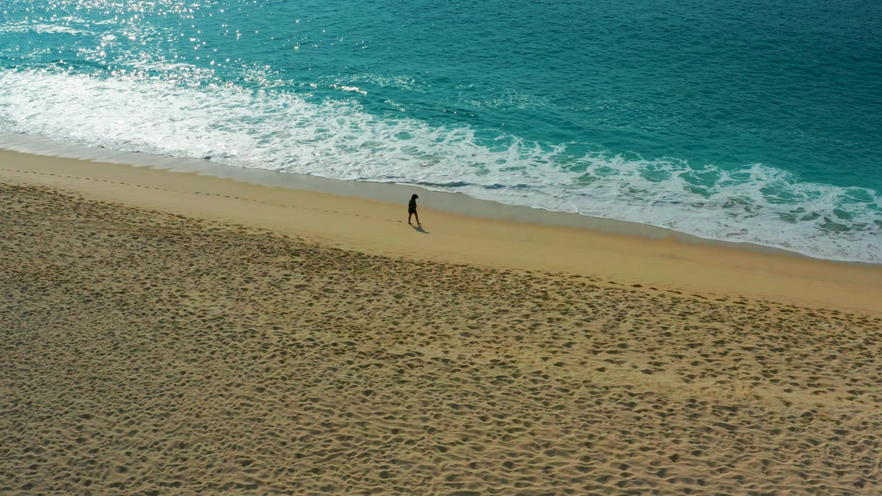 Aerial view in Desert Park Natural Reserve, Baja California Sur, showing one person walking along the shoreline as white surf breaks above a wide band of sand.
