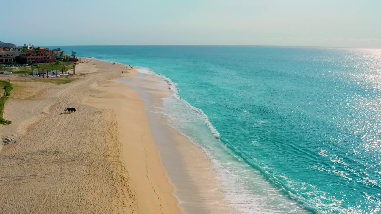 Aerial view over Desert Park Natural Reserve in Baja California Sur with the surf line running along the sand, sparkling turquoise water to the right, distant resort buildings to the left, and no visible people.
