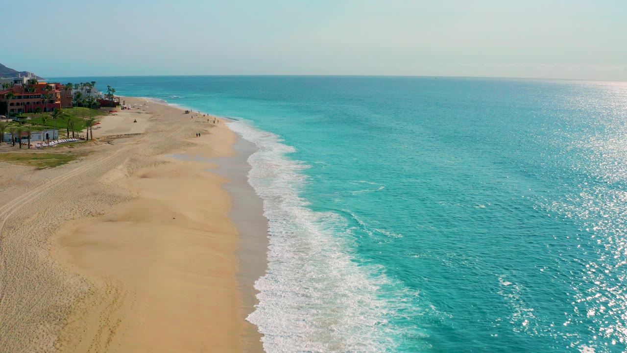 High aerial frame over Desert Park Natural Reserve in Baja California Sur, with turquoise ocean on the right, foamy surf along the sand, red resort buildings and palms on the left, and no visible people.