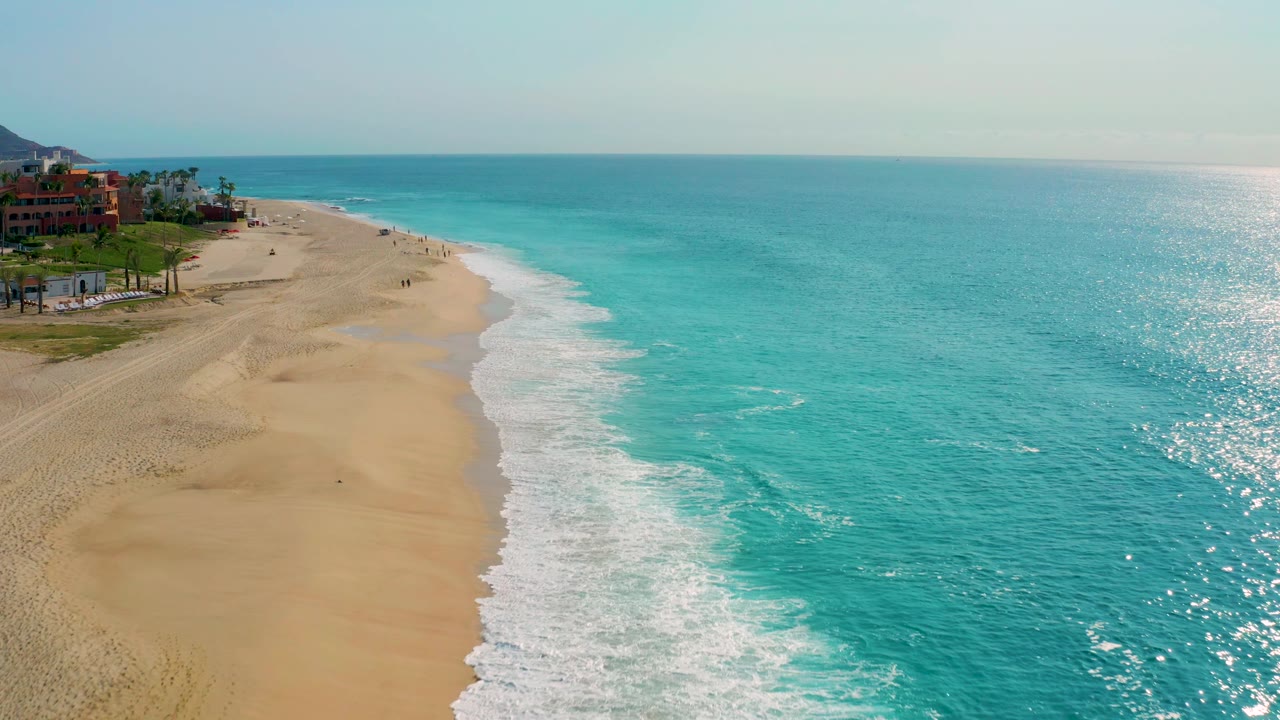 Wide aerial frame from Desert Park Natural Reserve showing sparkling turquoise ocean across the right, pale sand and surf along the left, red resort structures near trees, a low mountain behind them, and no visible people.