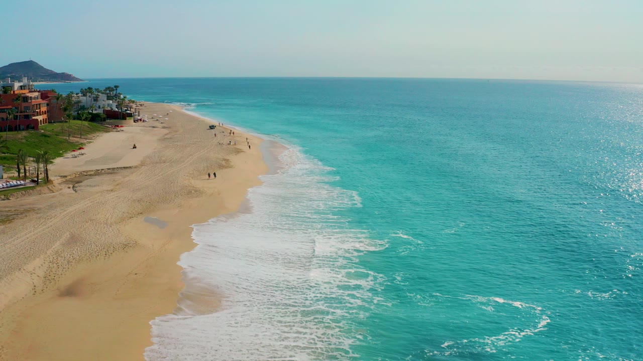High aerial view over Desert Park Natural Reserve in Baja California Sur, with turquoise water on the right, a wide sandy beach on the left, small distant walkers, shoreline buildings, palms, and a hill on the horizon.