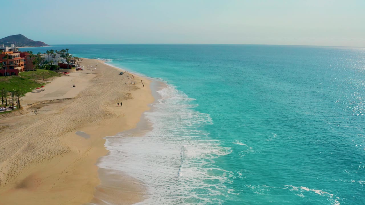 High-angle Baja California Sur coastline view with more open turquoise water to the right, a curving band of white surf, broad sand, tiny figures on the beach, buildings at far left, and a hill beyond the shoreline.
