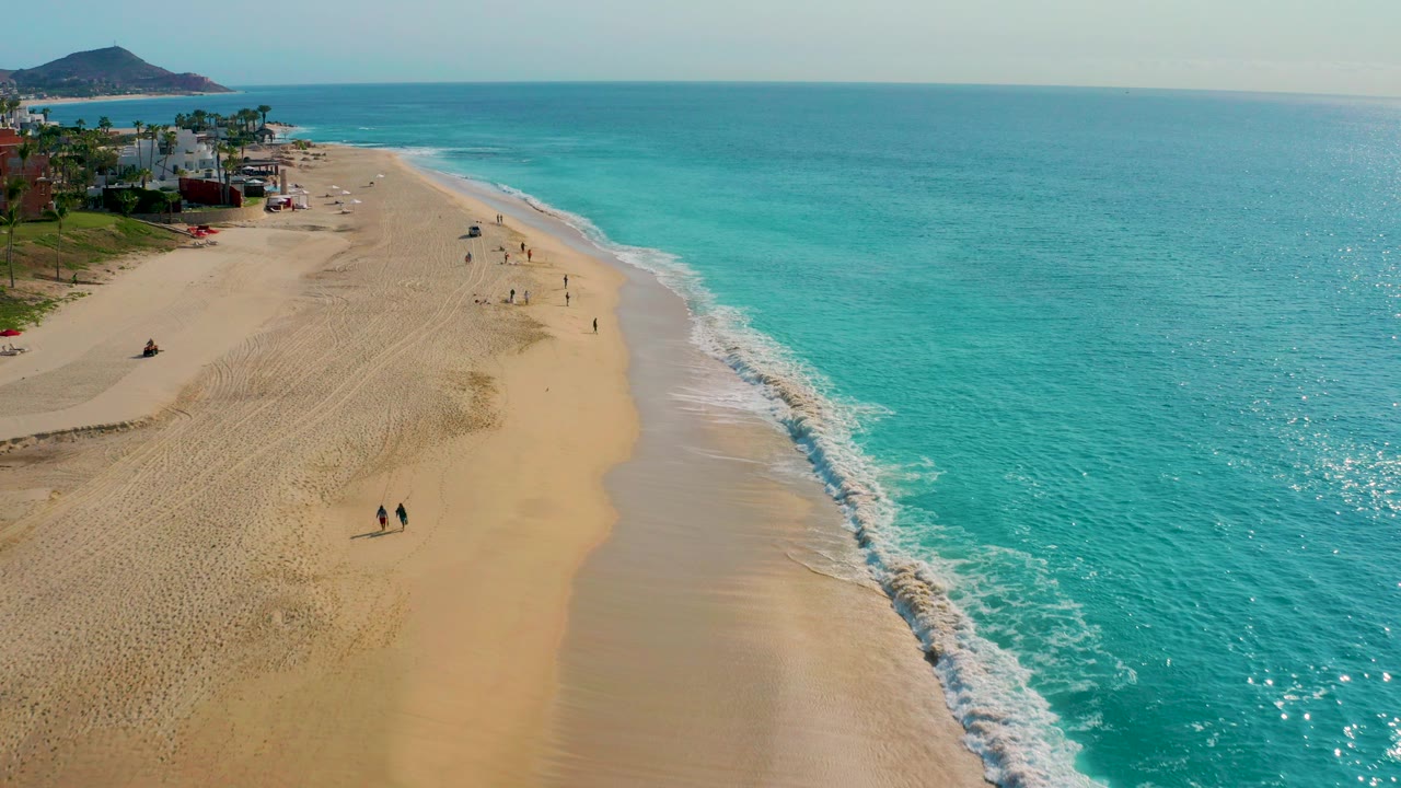 High aerial frame over Desert Park Natural Reserve in Baja California Sur, with tan beach at left, turquoise ocean at right, white surf, distant low-rise buildings, a hill, and small people walking on the sand.