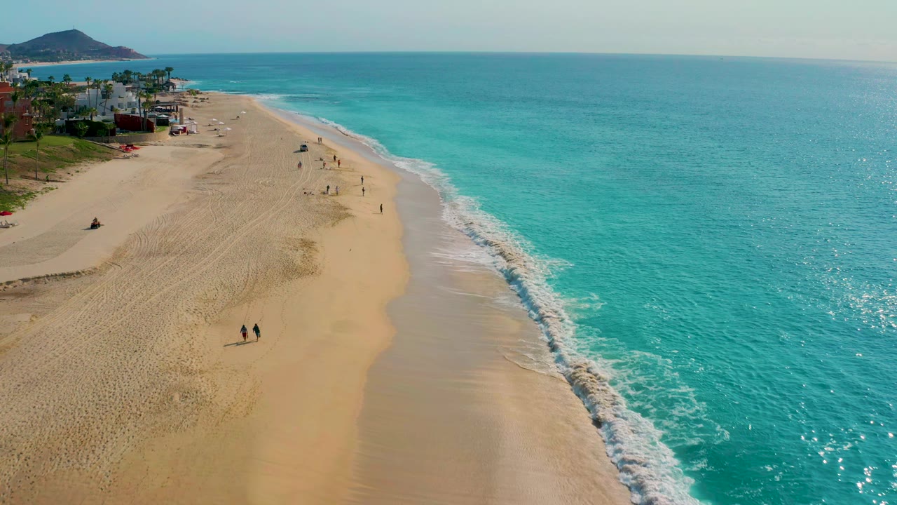Drone view down a sandy shoreline with turquoise water, surf foam, walkers, buildings, and a hill.
