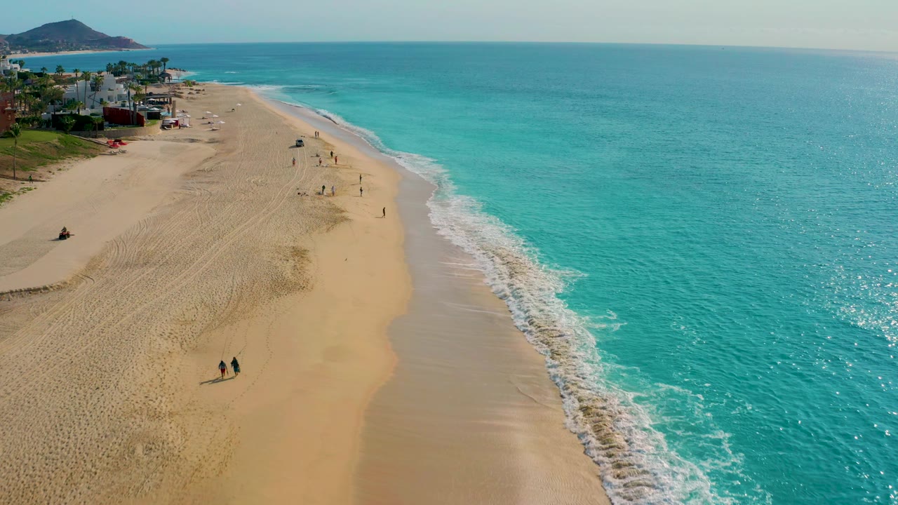 Wide drone frame from Baja California Sur with two walkers more prominent on the lower sand, small groups farther up the beach, turquoise water filling the right side, and the village edge near the top left.