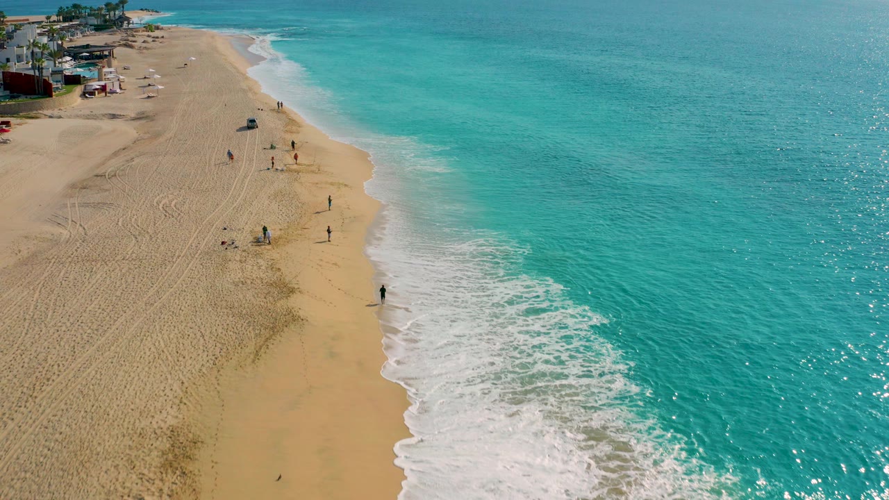 High-angle aerial frame from Desert Park Natural Reserve in Baja California Sur showing a wide sandy beach, turquoise ocean, white surf, and small visible people along the shoreline.