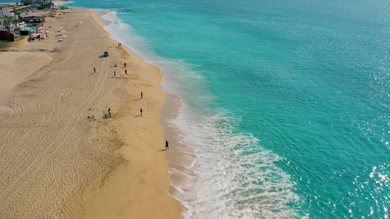 High-angle view of Desert Park Natural Reserve beach with turquoise sea taking most of the right side, a curved foam line, and scattered visible people on the sand.