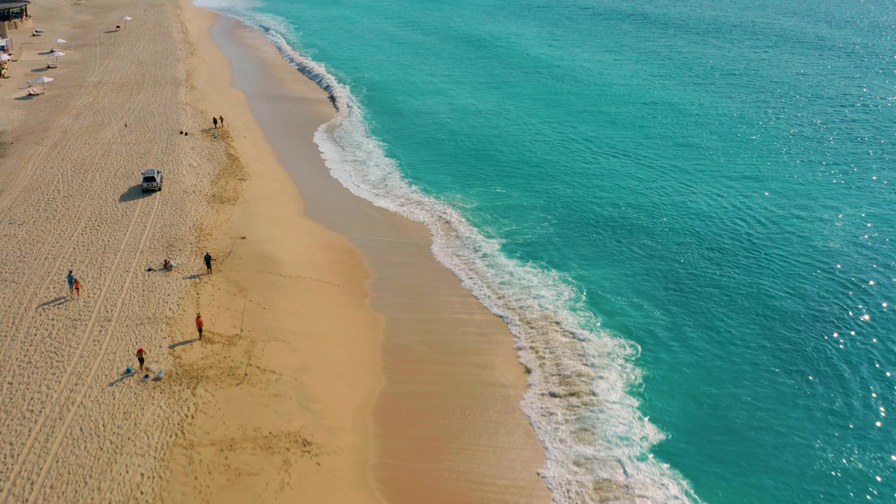 High-angle aerial over Desert Park Natural Reserve in Baja California Sur, Mexico, showing turquoise ocean on the right, foamy surf, golden sand, small beachgoers, umbrellas, and a silver vehicle near the upper beach.