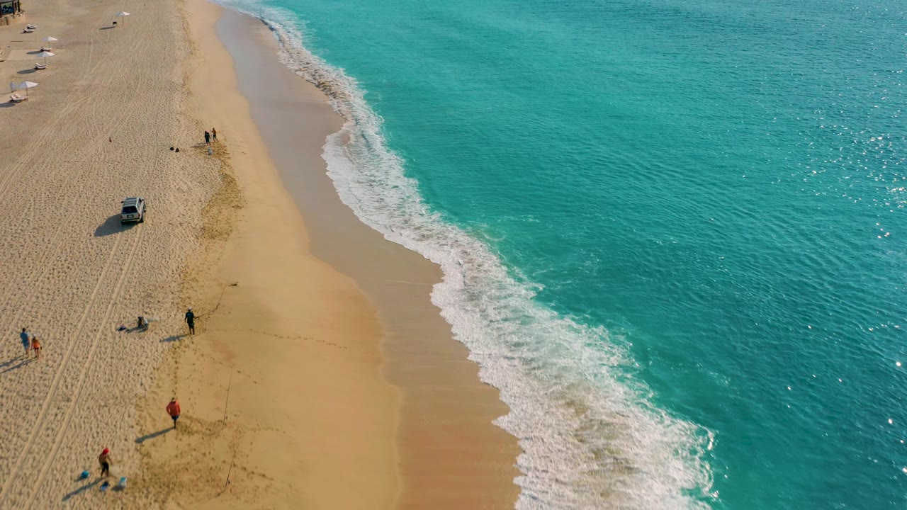 Drone view over Desert Park Natural Reserve in Baja California Sur, Mexico, where a silver vehicle sits farther inland from the wet shoreline, people stand near the sand tracks, and bright surf breaks along the right side.