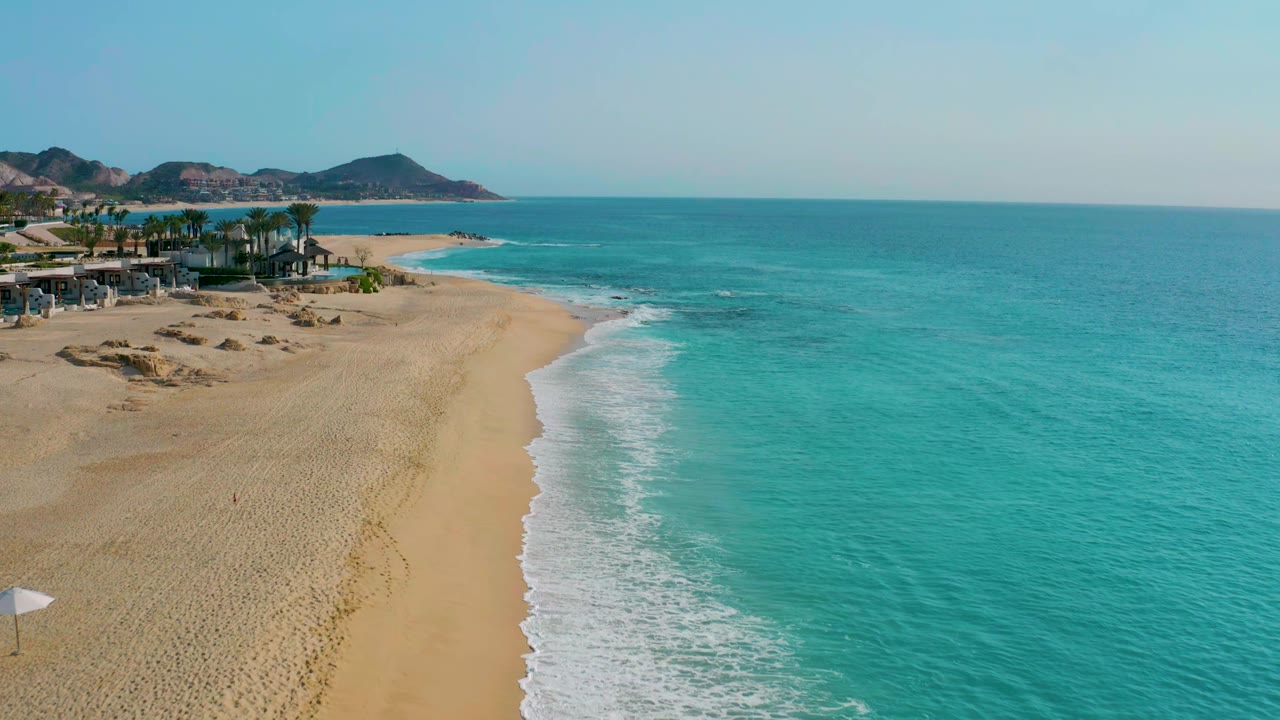 Drone view of the Baja California Sur coastline with the beach sweeping diagonally into turquoise water, palm-lined structures at left, mountains in the distance, and no visible people.