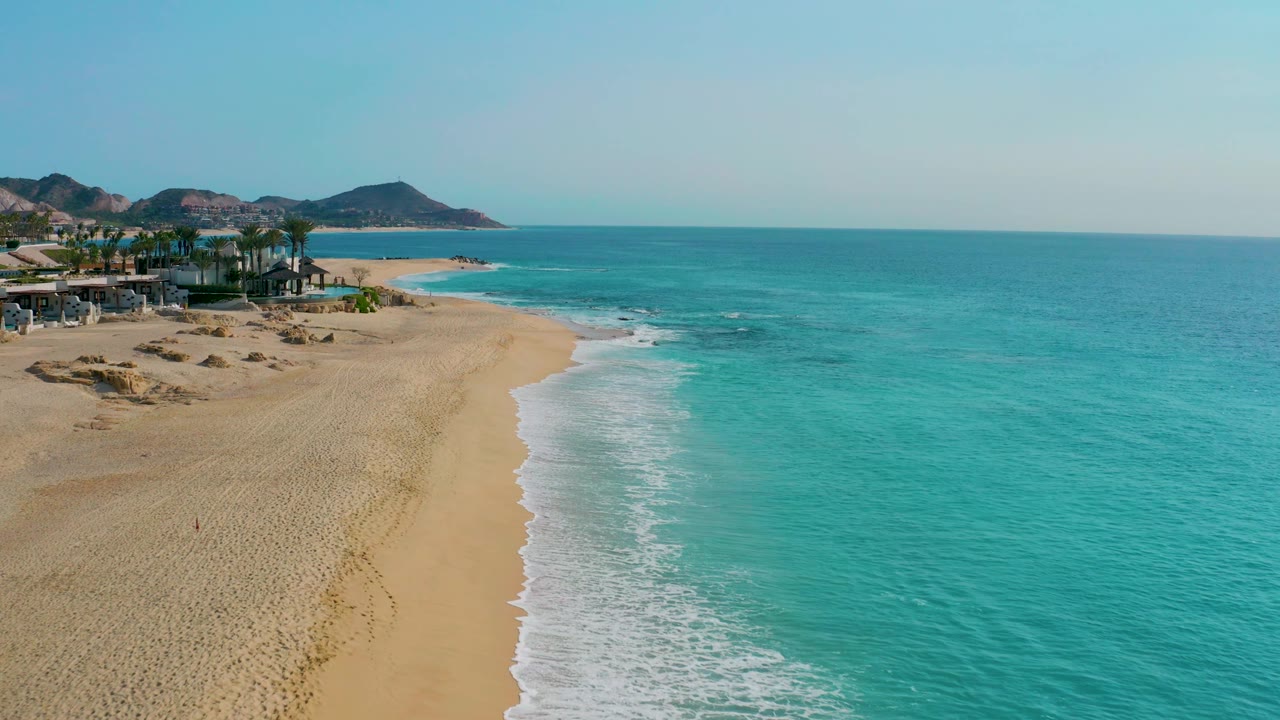 Aerial view along the Desert Park Natural Reserve shore with white foam tracing the sand, open ocean to the right, developed beachfront and palms to the left, distant mountains, and no visible people.
