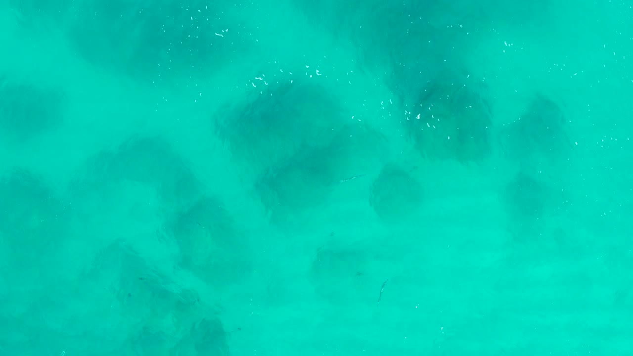 Overhead view of turquoise ocean surface in Desert Park Natural Reserve, Baja California Sur, with muted sand ripples and soft darker shapes below, no visible people.